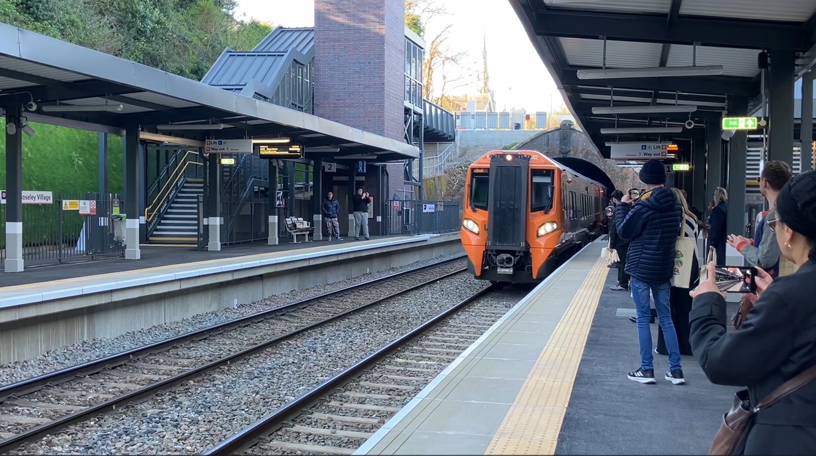 A railway station, with a train with orange coloured front facing camera pulling in. People on platform are holding up cameras and phones